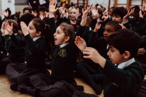 A group of enthusiastic schoolchildren, clad in green uniforms with school crests, are seated on the floor, actively participating in a group activity. Their expressions are animated and joyful as they raise their hands and clap, fully engaged in the interactive learning experience. The atmosphere is lively and collaborative, highlighting the children's enjoyment and involvement in the educational moment.