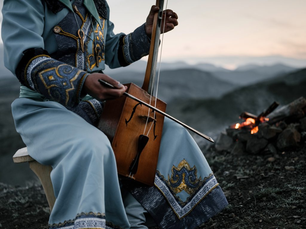 A person dressed in a traditional blue and gold embroidered outfit plays a stringed instrument, possibly a morin khuur, against a backdrop of a dusky sky and mountainous landscape. The individual is seated beside a small campfire, with the focus on their hands and the instrument, evoking a sense of cultural tradition and the serene ambiance of outdoor music.