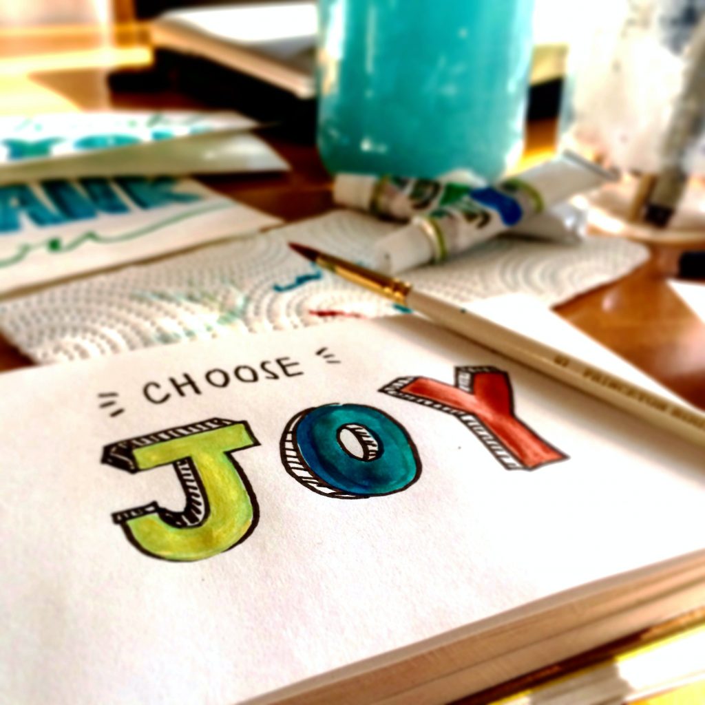 The image shows a close-up of a hand-drawn artwork with the phrase "CHOOSE JOY" in colorful, bold letters on a white paper. Surrounding the paper are art supplies, including paint tubes, brushes, and a jar of water, all indicative of an ongoing creative process. The focus on the inspiring message suggests a positive and artistic atmosphere.