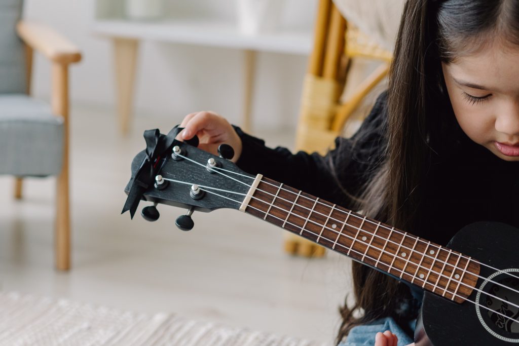 A young girl is focused on tuning a black ukulele, her delicate fingers adjusting the tuning pegs. The close-up captures her concentration and the fine details of the instrument, set against a softly blurred background of a cozy, well-lit room.