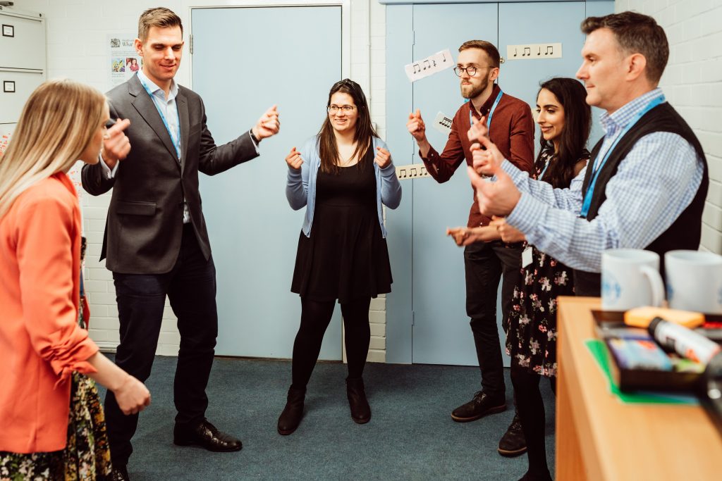 A group of six adults, likely educators, are engaged in a lively activity in a classroom setting, with some clapping and others gesturing as if conducting music. They are informally dressed, some with lanyards, suggesting a collaborative workshop or training session. The atmosphere is one of focus and enjoyment, with musical notation decorations on the wall adding to the educational ambiance.