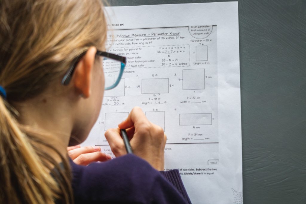 A student is closely examining a math worksheet focused on calculating perimeters. The worksheet includes various problems where the perimeter is given, and the student must find the measure of the unknown side of different shapes. The student appears to be engaged in solving the problems, with a pen in hand ready to write down the answers.