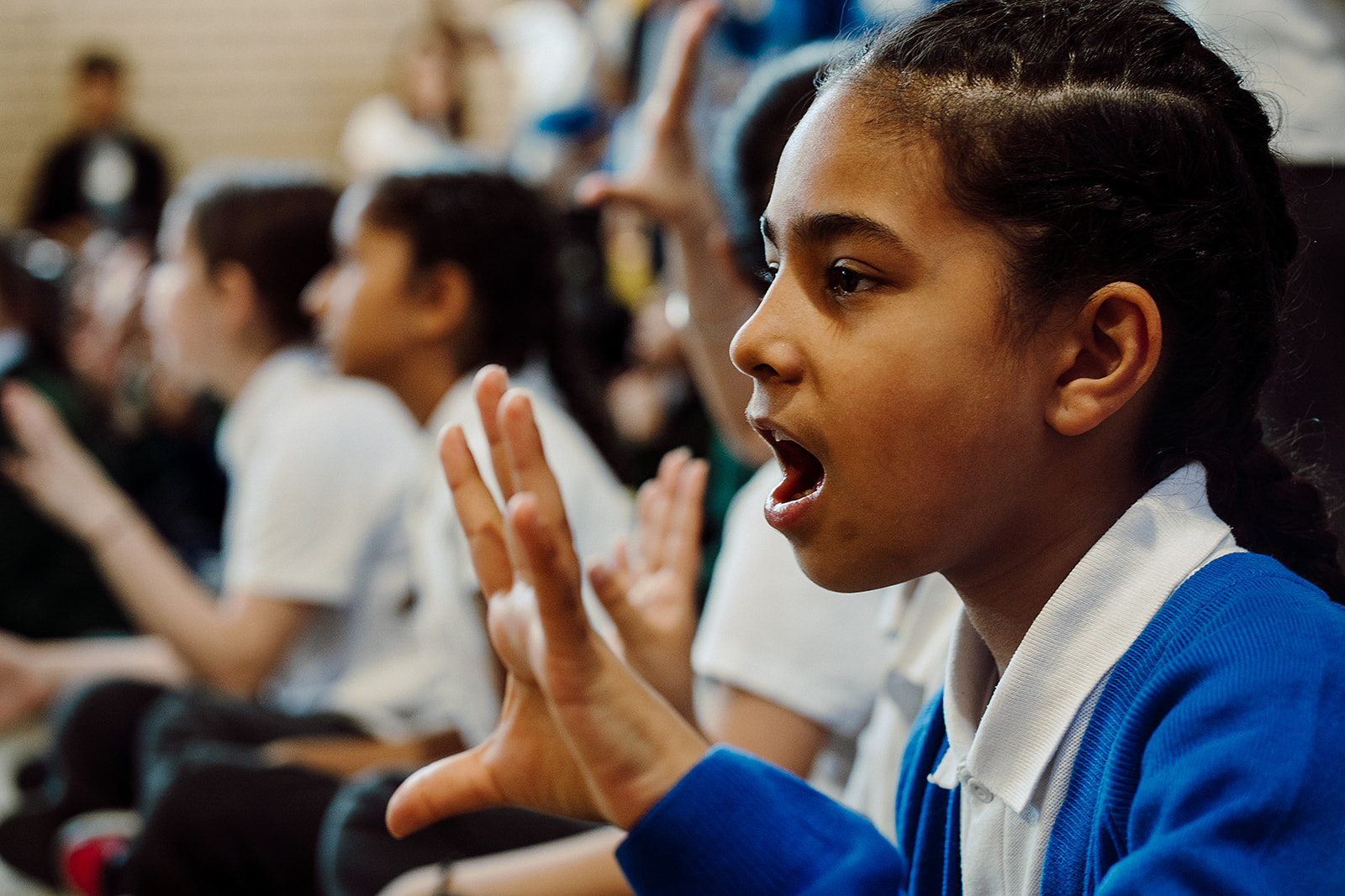 A young student in a white shirt and blue sweater is captured mid-song, her expression one of focus and joy as she participates in a group singing session. Her hands are expressive, accentuating the emotion of the performance, while blurred figures in the background suggest a communal atmosphere of shared musical experience.