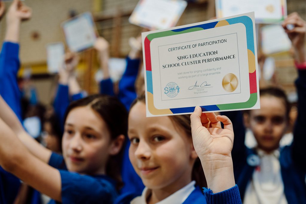 Proud students in blue uniforms hold up their Sing Education certificates of participation, celebrating their achievements in a school cluster performance. The focus is on a certificate that commends singing confidently and working as part of a large ensemble, reflecting the sense of accomplishment and teamwork fostered through the program.