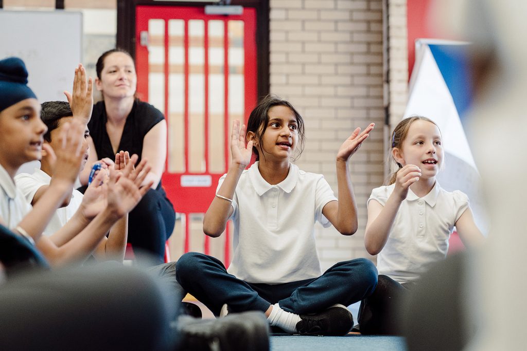 A group of primary school children are seated on the floor, actively participating in a classroom activity with raised hands and animated expressions. A teacher, slightly out of focus in the background, observes with a smile, indicating a positive and engaging learning environment. The children's attention is directed towards something or someone not visible in the frame, suggesting interactive learning.