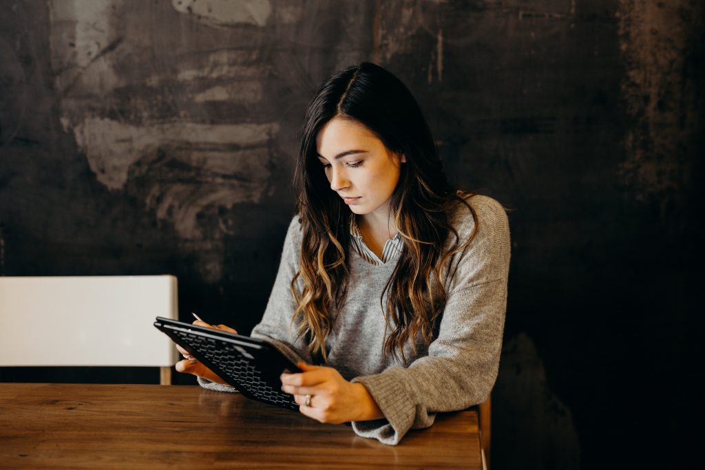 A focused woman sits at a wooden table, engrossed in reading or working on a tablet. She's dressed in a cozy grey sweater over a collared shirt, suggesting a casual, professional or educational setting. The background features a textured dark wall, adding a rustic, contemporary ambiance to the scene.
