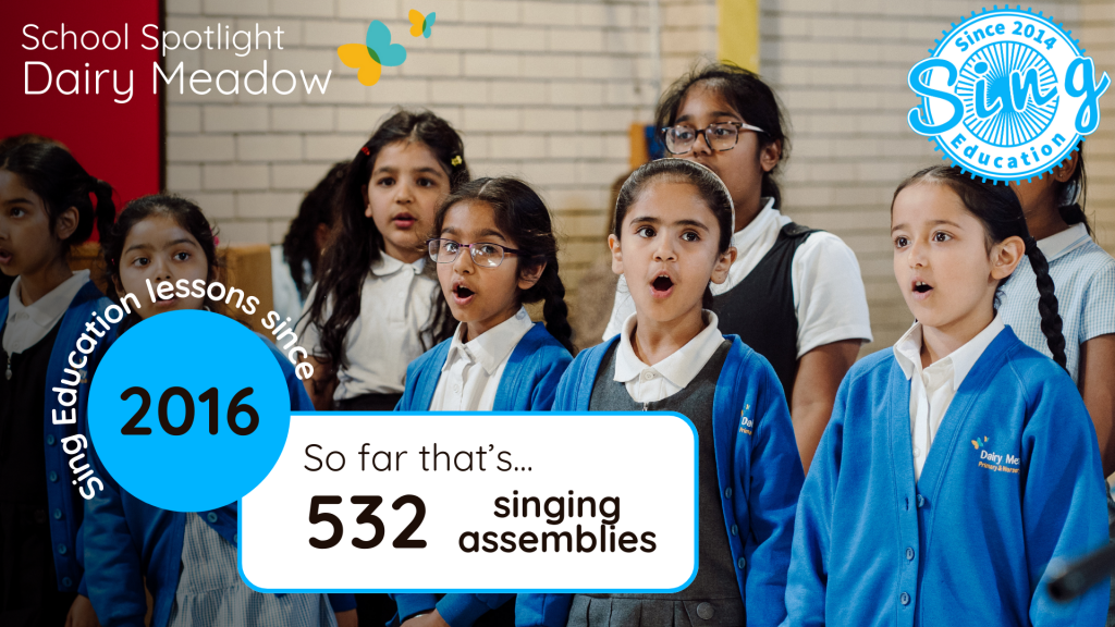 A group of enthusiastic young students in school uniforms are engaged in a singing assembly, with expressions of focus and enjoyment as they participate in the musical activity. The overlaying graphics highlight the school's name, Dairy Meadow, and celebrate the number of singing assemblies conducted by Sing Education since 2016, indicating a total of 532 assemblies. The image conveys a sense of community and the joy of collective music-making in an educational setting.