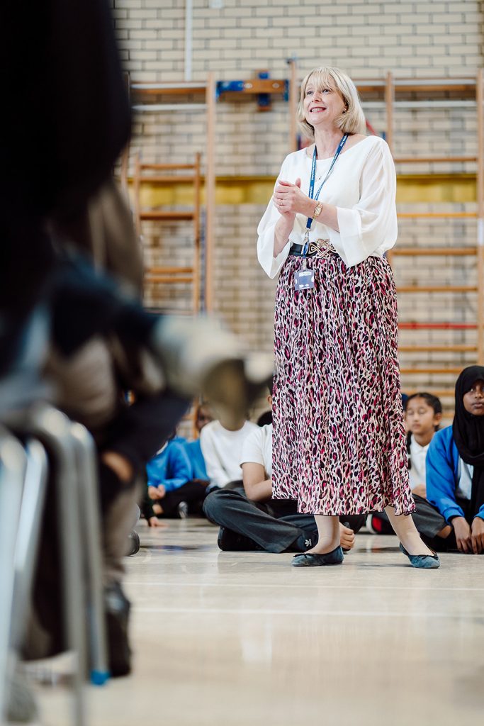 A smiling educator stands confidently in a school gym, engaging with a circle of seated students who watch attentively. Her hands are clasped together, and her expression conveys enthusiasm, suggesting an interactive and positive learning environment.
