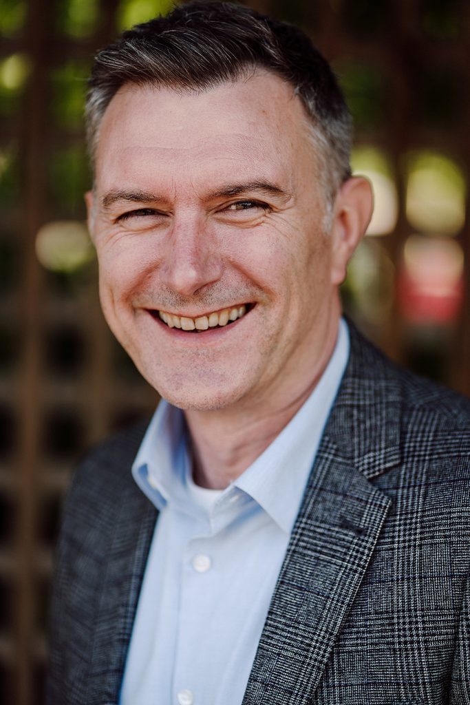 A smiling man with a neatly combed hairstyle is captured in a close-up portrait. He wears a patterned blazer over a light blue shirt, exuding a professional yet approachable demeanor. The background is softly blurred, highlighting his cheerful expression.