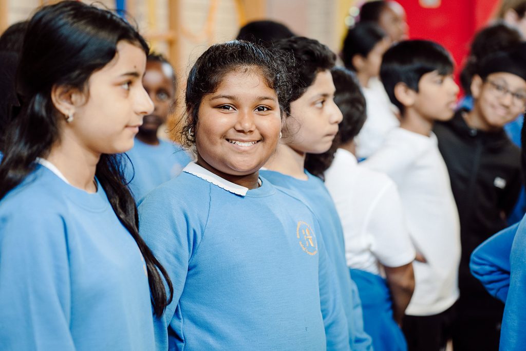 A group of schoolchildren in uniform, with a focus on a smiling girl in the foreground, captures a moment of joyful engagement among students. The children's attentive expressions suggest a shared experience, likely in a classroom or assembly setting, fostering a sense of community and learning.