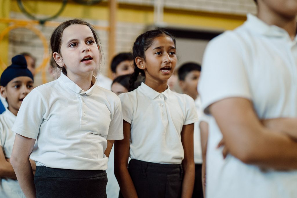A group of schoolchildren, wearing white polo shirts, are gathered together, attentively participating in a group activity. Two girls in the foreground appear engaged and focused, possibly singing or responding to a teacher's instruction, as they look slightly upwards with mouths open as if in mid-speech or song. The atmosphere suggests a lively and interactive learning environment.