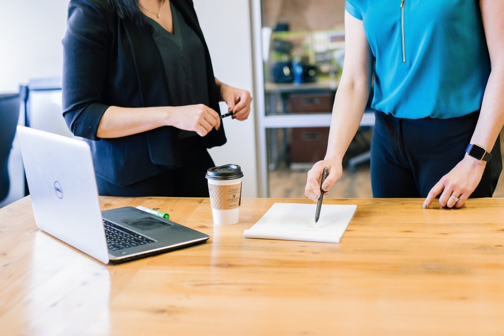 Two individuals stand at a wooden table in an office setting, collaborating on a task. One person, wearing a black blazer, holds a pen while the other, in a blue top, writes on a paper. A laptop, a disposable coffee cup, and a smartphone are also on the table, suggesting a professional meeting or work session.