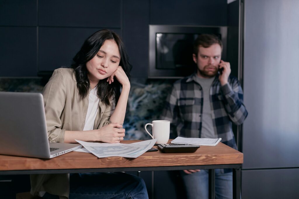 A woman sits at a kitchen table, appearing contemplative or tired, with papers and a laptop in front of her, while a man stands behind her, talking on the phone with a serious expression. The setting suggests a moment of domestic life, possibly involving work or personal matters, with a mug and calculator also on the table, indicating a blend of household and professional or financial activities.