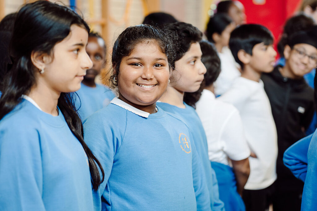 A group of schoolchildren in uniforms stands together, with a focus on a smiling girl in a blue sweater at the forefront, exuding confidence and happiness among her peers. The blurred background suggests a bustling school environment, highlighting the sense of community and camaraderie among the students.