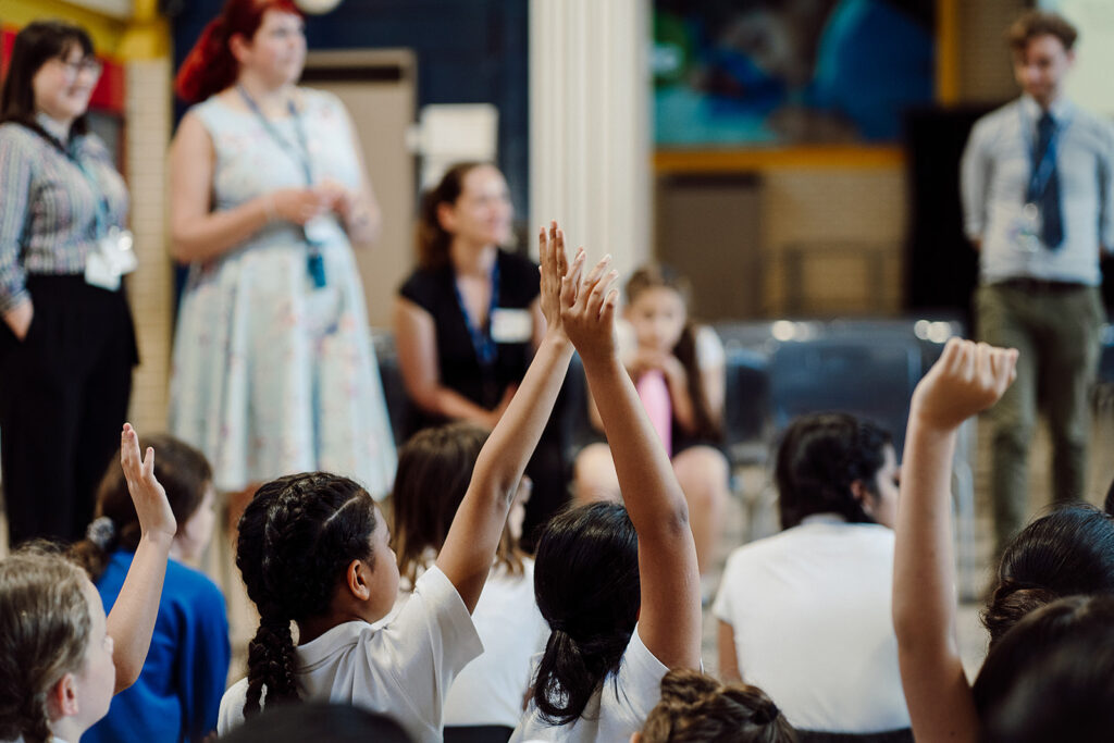 Children are sitting on the floor with their backs to the camera. Some children have their hands raised to answer a question. In the background a group of teachers is watching the lesson