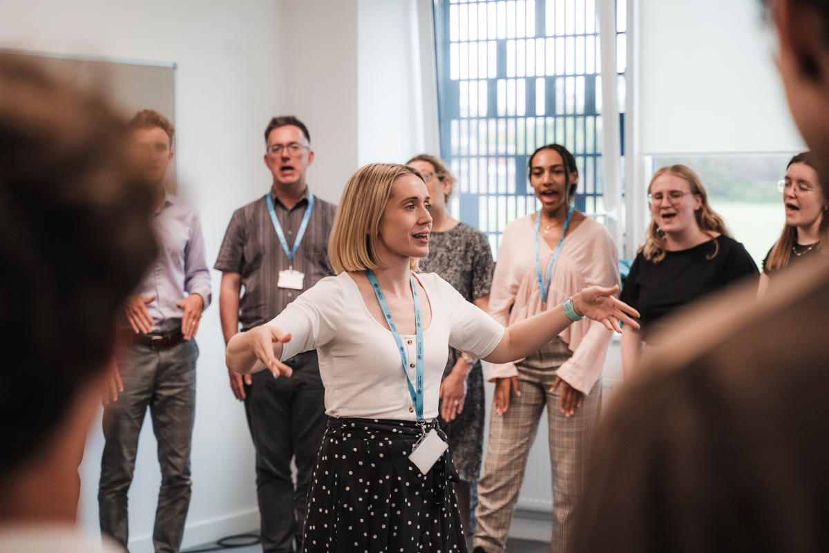 A woman with a white shirt is standing in the middle of a circle of other teachers. She has a realxed and confident expression as she trains the other people in the room