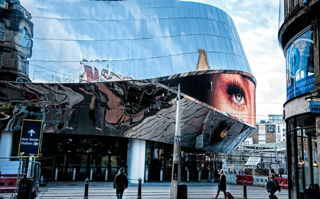 People walking near a glass building in Birmingham. In the bottom left corner there is a sign pointing to New Street Station and the reflections of the city can be seen in the glass windows of the building.