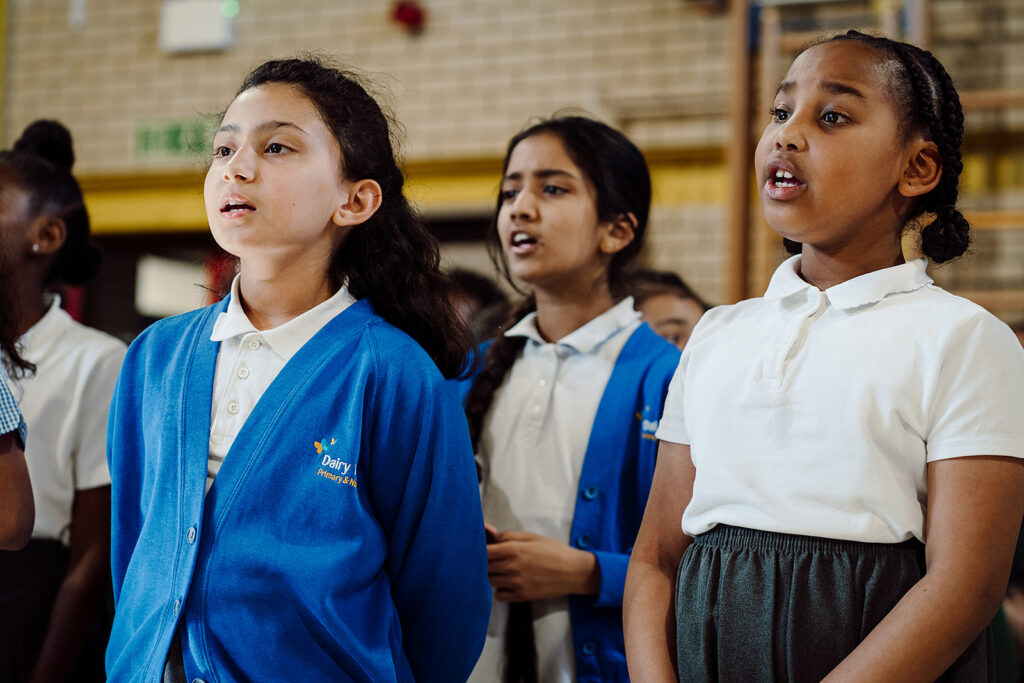 Children wearing a blue uniform with a white polo shirt are singing and looking off camera to the left. They are performing in a singing concert
