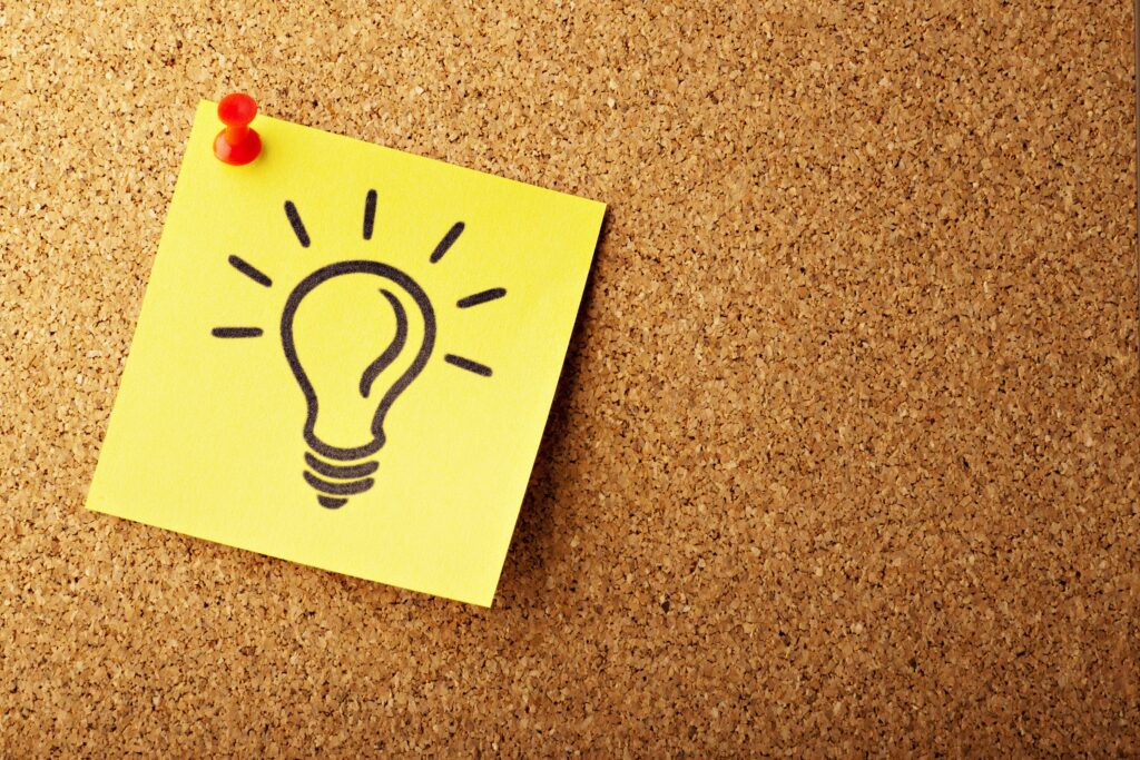 A post it note with a drawing of a lightbulb is pinned to a cork board