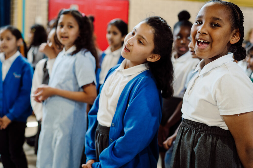 A row of children wearing a blue uniform are smiling and singing. One child is looking directly at the camera and smiling