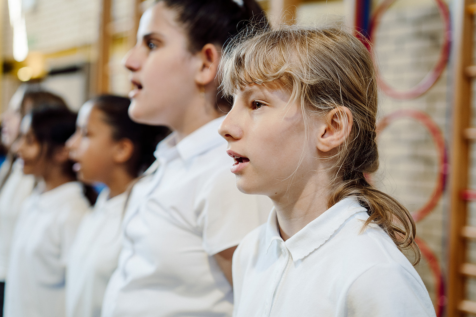 The photo is taken looking down a line of children singing. They are wearing white polo shirts for their school uniform