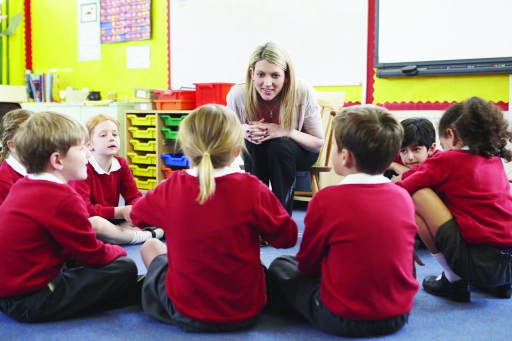 A class teacher is looking at children sitting on the floor. They are wearing a red school uniform