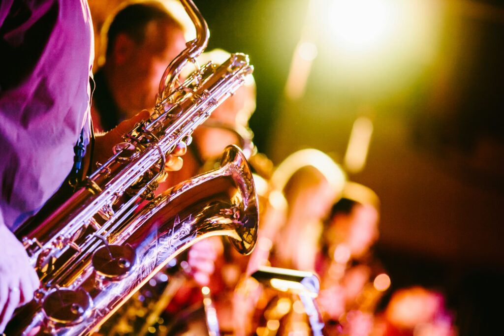 A saxophone is in the foreground. In the background other instruments are being played. The lights are bold and colourful showing us they are performing on stage
