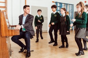 A music instructor plays the piano, capturing the attention of a group of uniformed schoolchildren who are actively participating in a music lesson, with expressions of focus and enjoyment as they engage in the learning experience.